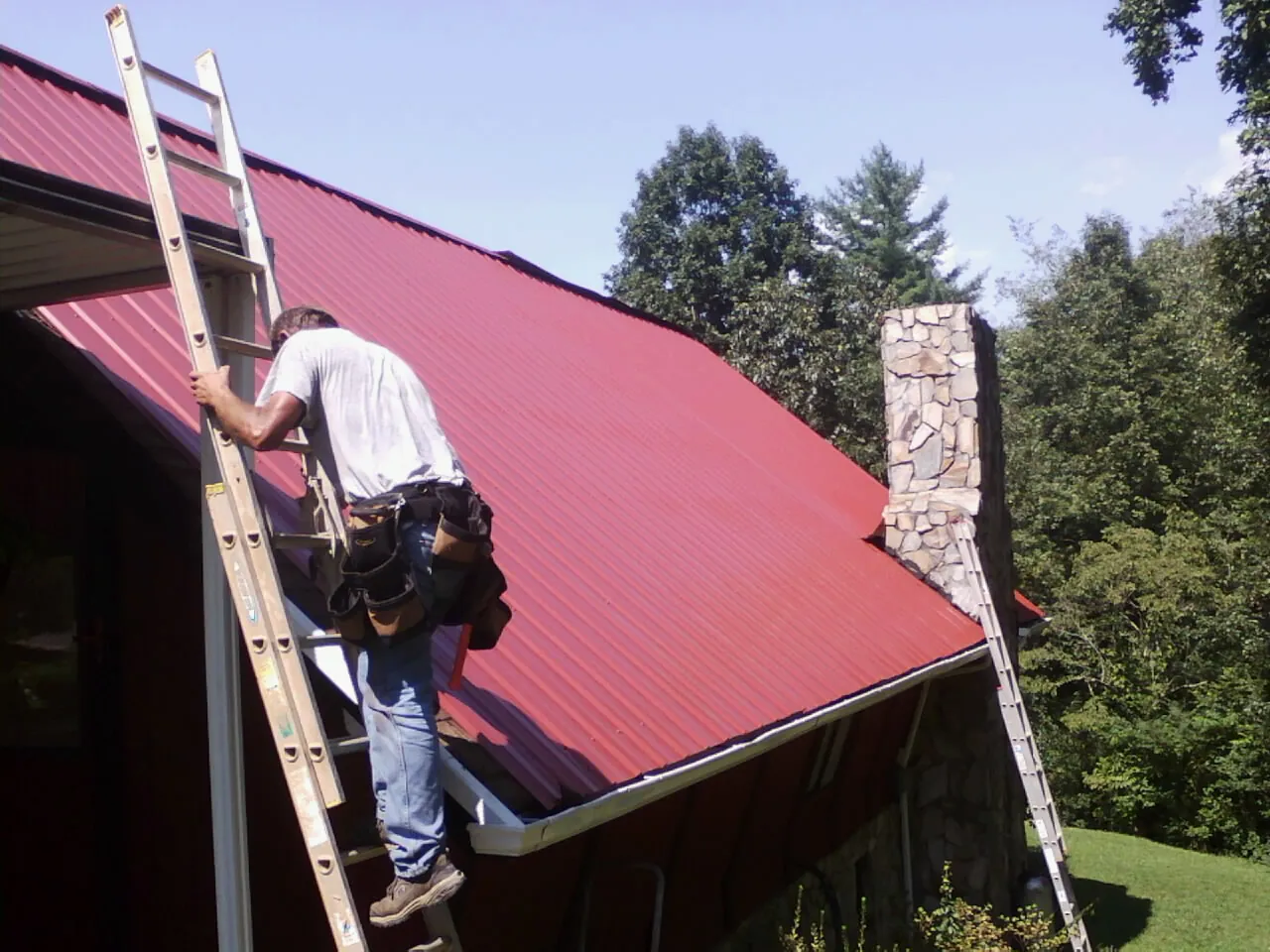 Roofer on a ladder beside a red metal roof and stone chimney — Soffit & Fascia Repair by Roofing Brothers in Paupack